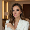 A woman with long brown hair wearing a white blazer, posing in a warmly lit indoor setting.